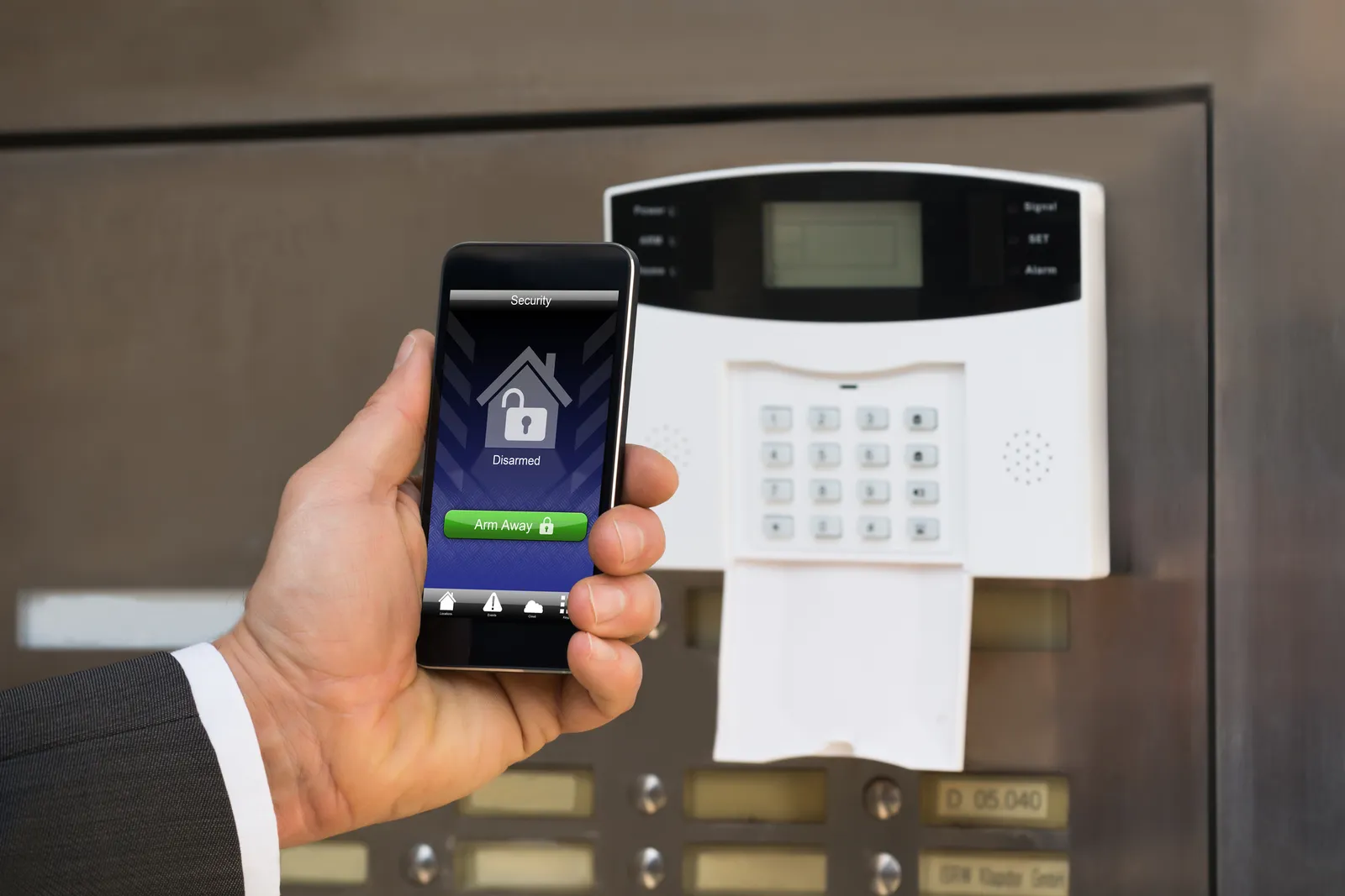 Empresario sosteniendo smartphone con app de seguridad frente a panel de alarma físico