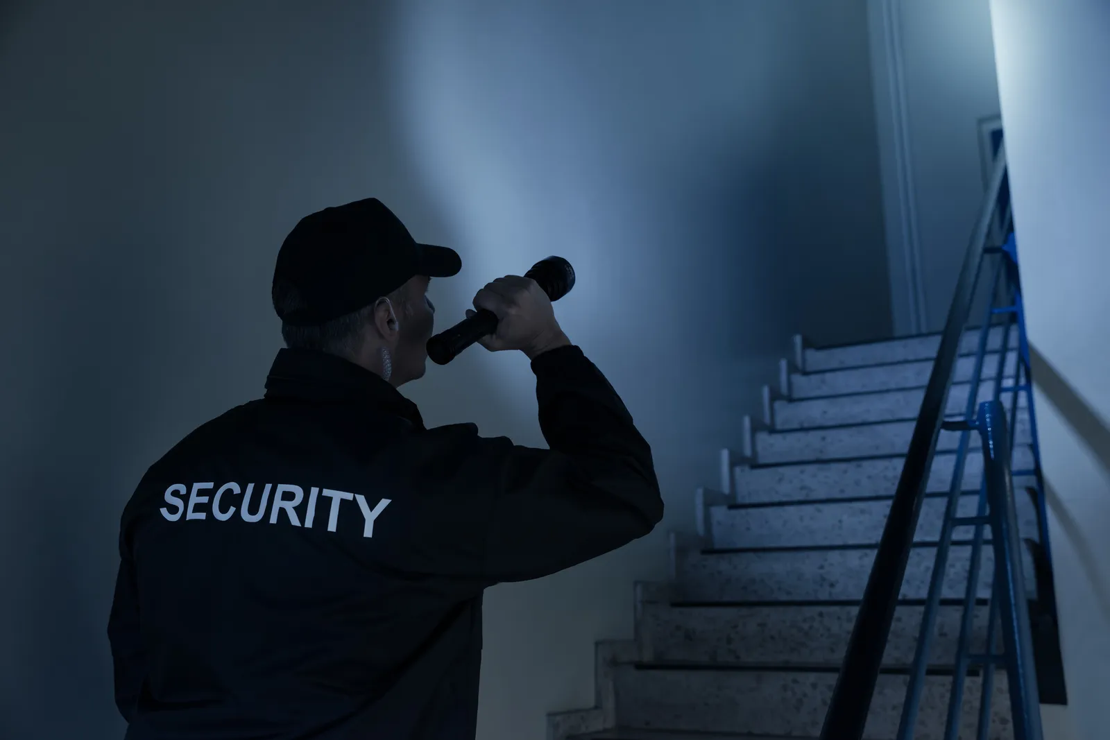Guardia de seguridad de espaldas con linterna iluminando escalera en ronda nocturna