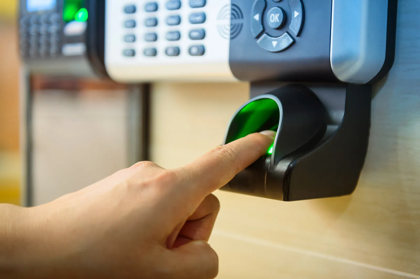 Lector biométrico de huella digital con sensor verde montado en pared corporativa con persona acercando el dedo