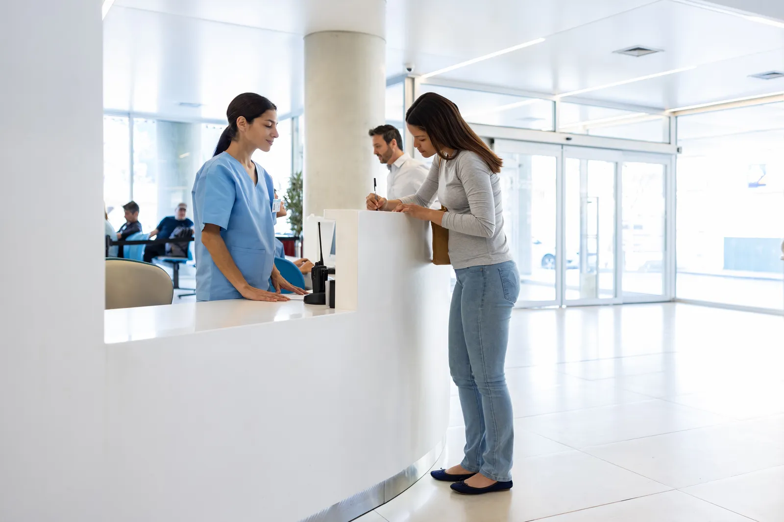 Recepción de una clínica privada con una paciente completando el formulario de admisión y la sala de espera al fondo
