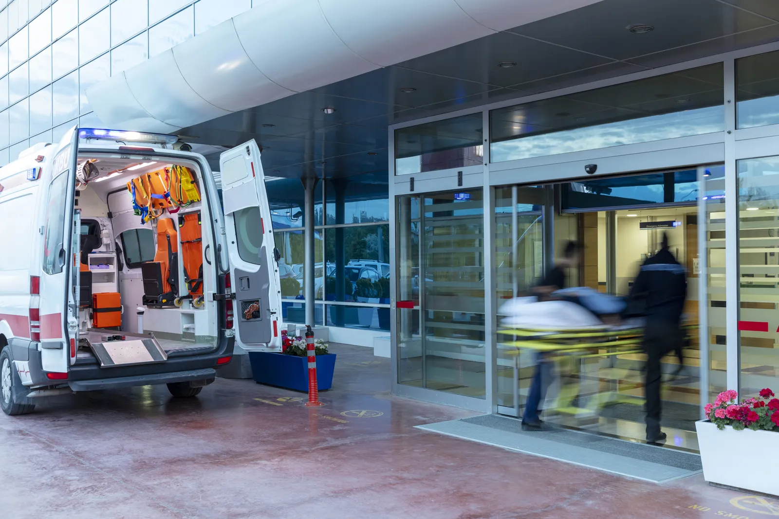Ambulancia con las puertas abiertas en la rampa de emergencias de un hospital mientras ingresan a un paciente en camilla