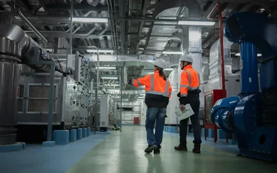 Dos operarios con chaleco y casco recorriendo la sala de máquinas de una planta industrial