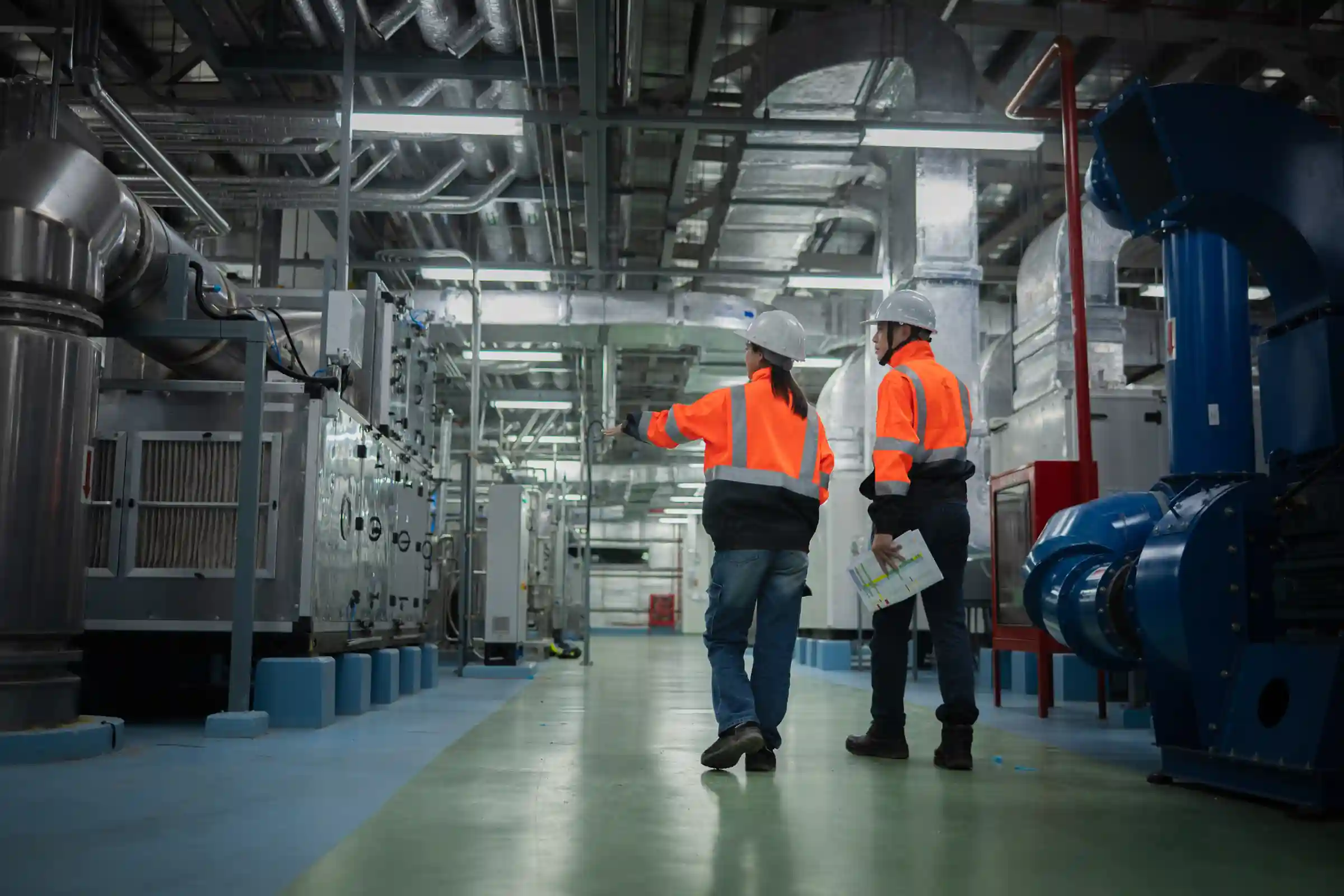 Dos operarios con chaleco y casco recorriendo la sala de máquinas de una planta industrial